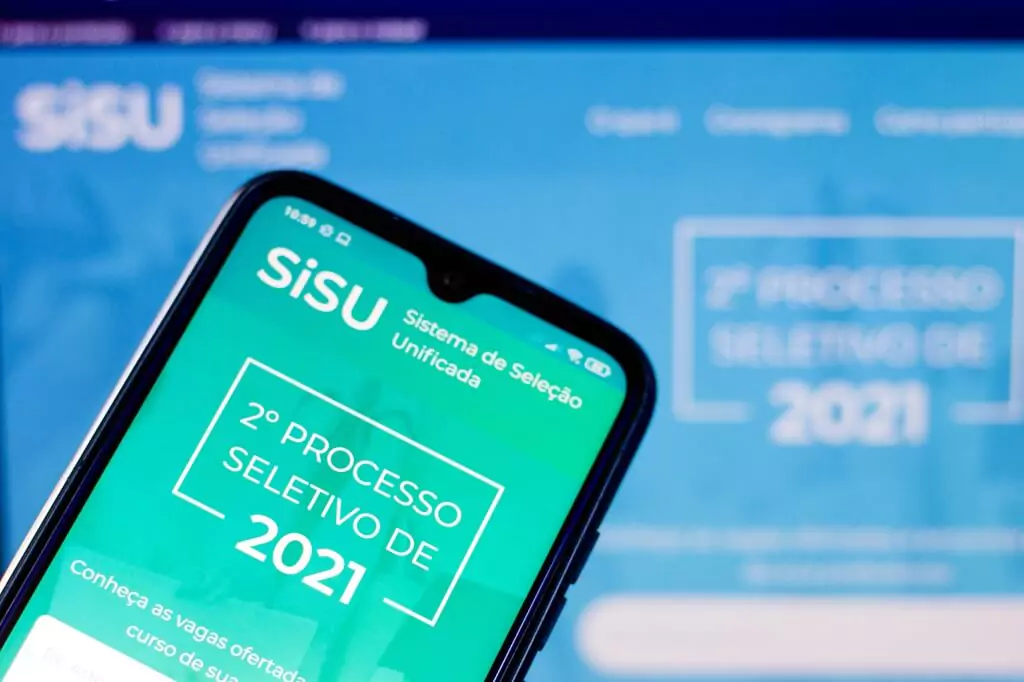 Celular mostrando app do Sisu com tela do site ao fundo em computador